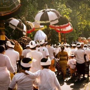 Un grande gruppo di persone in abiti tradizionali bianchi sfila in processione, portando grandi ombrelli cerimoniali lungo una strada alberata e illuminata dal sole.