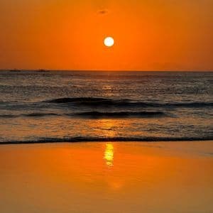 Il sole tramonta sull'oceano, proiettando un bagliore arancione nel cielo e sull'acqua. Il suo riflesso brilla sulla sabbia umida della spiaggia.
