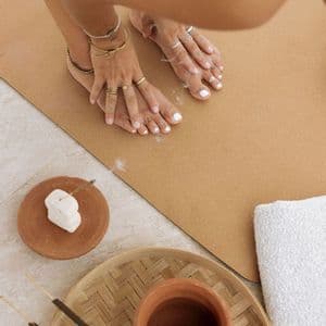 Una persona in posizione yoga di piegamento in avanti tocca i piedi su un tappetino marrone, con incenso che brucia e un asciugamano in primo piano.