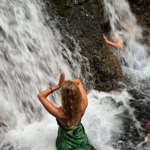 Una donna di un viaggio di gruppo WeRoad, ripresa di spalle, in posa di preghiera nell'acqua impetuosa di una cascata.