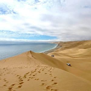 Vaste dune di sabbia digradano verso l'oceano, con due veicoli 4x4 bianchi che percorrono la linea costiera.