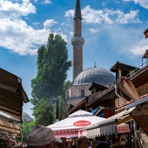 Una folla di persone si muove in una vivace strada del mercato, con una moschea di pietra e il suo minareto che si stagliano contro un cielo blu e nuvoloso.