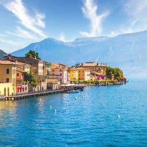 Edifici colorati di una cittadina di lago si specchiano nell'acqua azzurra e brillante, con grandi montagne sullo sfondo sotto un cielo soleggiato.