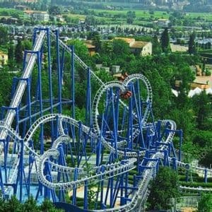Vista aerea di imponenti montagne russe blu e bianche, con numerosi anelli e avvitamenti, immerse nel verde lussureggiante di un parco divertimenti.