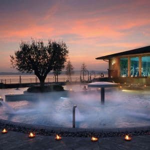 Una piscina termale esterna fumante in un resort benessere, circondata da candele accese e affacciata su uno specchio d'acqua al tramonto.