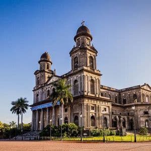 Una grande cattedrale in pietra, segnata dal tempo, con due torri si erge in una piazza soleggiata accanto a palme sotto un cielo sereno.