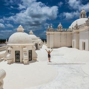 Una donna con le braccia alzate è in piedi sul vasto tetto bianco di un edificio con cupole multiple sotto un cielo blu nuvoloso.