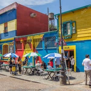 Le persone siedono ai tavoli sotto gli ombrelloni fuori da edifici dai colori vivaci su un angolo di strada soleggiato.