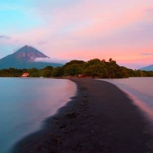 Una lingua di sabbia nera si curva in un lago calmo, con un grande vulcano visibile sullo sfondo sotto un cielo colorato al tramonto.