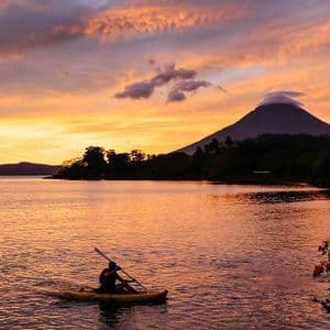 Una persona in kayak giallo su un lago al tramonto vibrante, con un grande vulcano stagliato sullo sfondo.