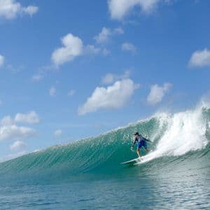 Un surfista in pantaloncini blu cavalca una grande onda turchese sotto un cielo parzialmente nuvoloso.