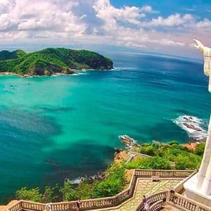 Un'alta statua bianca di Gesù si erge su una scogliera, dominando una baia turchese con barche e colline verdi.