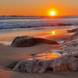 Il sole tramonta sull'oceano, proiettando un bagliore arancione sulle onde, sulla sabbia bagnata e sulle grandi rocce in primo piano.