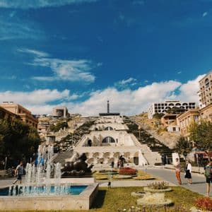 Eine monumentale terrassierte Steintreppe an einem Hang, gesehen von einem Park mit Springbrunnen, Skulpturen und Gärten unter blauem Himmel.