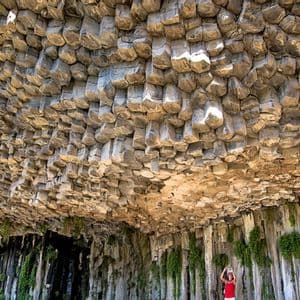 Eine Person in einem roten Oberteil steht und blickt auf eine riesige Decke aus sechseckigen Basaltsäulen in einer Felshöhle.