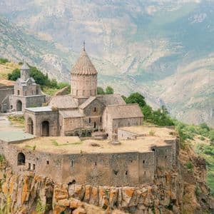 Ein historischer steinerner Klosterkomplex thront auf einem felsigen Kliff mit Blick auf ein weites, bergiges Tal.