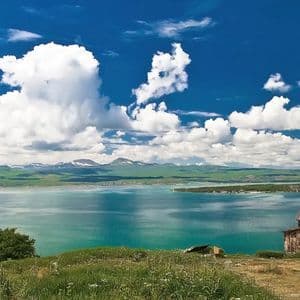 Eine alte Steinkirche mit kegelförmigem Dach steht auf einem grasbewachsenen Hügel mit Blick auf einen türkisfarbenen See und ferne schneebedeckte Berge.