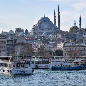 Ferries sail on the water before a dense cityscape, with a large mosque featuring a central dome and tall minarets on a hill.