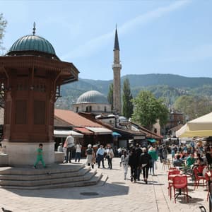 Une grande fontaine en bois se dresse sur une place de ville animée, avec une mosquée en arrière-plan et des gens se promenant parmi les cafés en plein air.