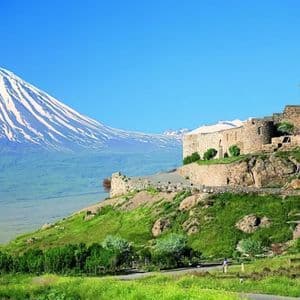 Ein Steinkloster auf einem grünen Hügel mit großem schneebedecktem Berg im Hintergrund unter klarem blauem Himmel.