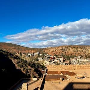 Un pueblo con construcciones de adobe se asienta en una ladera soleada de tono rojizo bajo un cielo azul brillante con nubes dispersas.