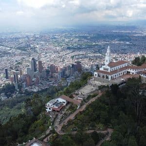Vue aérienne d'un sanctuaire blanc sur une montagne boisée dominant une ville moderne étendue sous un ciel nuageux.