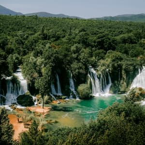 Vue panoramique de multiples cascades se déversant dans un bassin turquoise, entouré d'une forêt verte dense avec des collines en arrière-plan.
