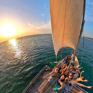 Un groupe WeRoad prend une photo en plongée sur un voilier en bois, naviguant sur l'océan au coucher du soleil.