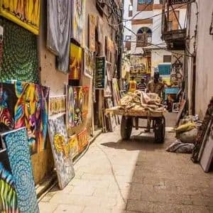 A narrow, sunlit alleyway market with colorful paintings of zebras and other animals displayed against the walls for sale.