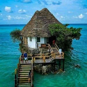 Un piccolo edificio con tetto di paglia sorge su una roccia nell'oceano turchese, con persone su una passerella di legno che vi conduce.