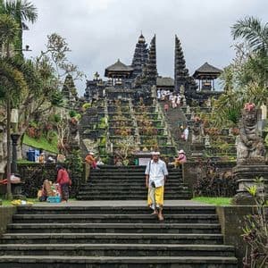 Ein Mann in weißem Hemd und gelbem Sarong schreitet die Steinstufen eines reich verzierten, mehrstufigen Tempels hinab, der von Gärten umgeben ist.