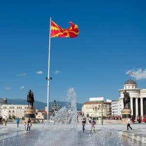 Die rot-gelbe Flagge Nordmazedoniens weht an einem hohen Fahnenmast auf einem belebten Stadtplatz mit Brunnen und Statuen unter einem klaren blauen Himmel.