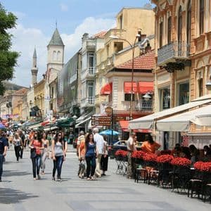 An einem sonnigen Tag schlendern Menschen durch eine belebte Fußgängerzone, gesäumt von Geschäften und Cafés mit Außenterrassen.