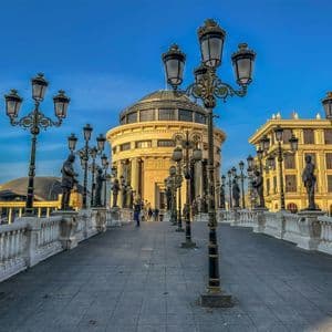 Eine breite Steinbrücke, gesäumt von zahlreichen Bronzestatuen und verzierten Laternen, führt zu einem prächtigen klassischen Gebäude unter blauem Himmel.