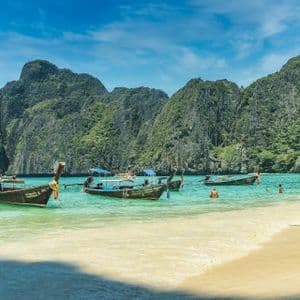 Plusieurs bateaux à longue queue sont amarrés dans une baie turquoise où des gens nagent, avec en arrière-plan de grandes falaises calcaires recouvertes de verdure.