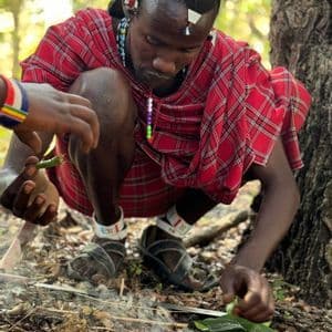 Un uomo con abiti tradizionali Maasai rossi è accovacciato sul suolo della foresta, intento a curare un piccolo fuoco fumante con foglie.