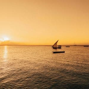 Un tradizionale veliero dhow e altre barche sull'oceano durante un tramonto dorato, con il sole che si riflette sull'acqua.