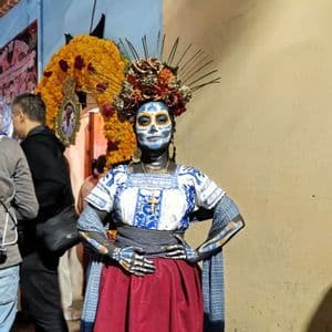 Eine Person in einem Catrina-Kostüm mit Totenkopf-Schminke und Blumenkopfschmuck posiert mit den Händen in den Hüften.