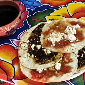 Ein Teller kleiner Maistortillas, garniert mit roten und dunklen Saucen und zerbröseltem Käse, neben einem Tonbecher auf einer bunten Blumentischdecke.