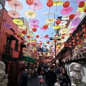 Bunte Papierschirme und rote Laternen hängen über einer belebten Stadtstraße, wo Menschen zwischen Gebäuden spazieren.