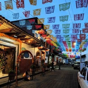 Bunte Papel Picado Banner hängen in der Abenddämmerung über einer engen Kopfsteinpflastergasse, die von mit Ringelblumengirlanden geschmückten Gebäuden gesäumt ist.