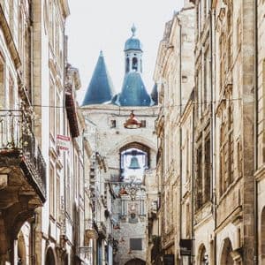 Una estrecha calle europea con edificios de piedra conduce a una caseta de vigilancia histórica con una gran campana visible bajo su arco.