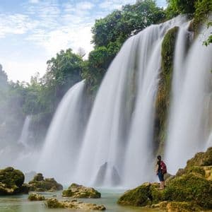 Une personne avec un sac à dos se tient sur des rochers moussus au pied d'une grande cascade, entourée d'un feuillage vert.