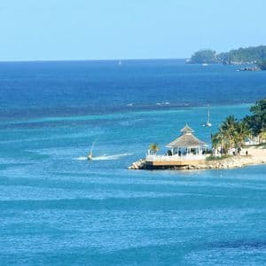 Un kiosque blanc sur une presqu'île domine une plage tropicale avec palmiers et bateaux sur la mer turquoise sous un ciel dégagé.