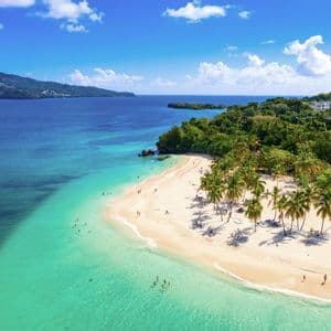 An aerial view of a tropical beach with people swimming in turquoise water and relaxing on white sand next to lush palm trees.
