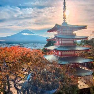 Una pagoda rossa giapponese sorge tra alberi dal fogliame autunnale, affacciata su una città con un grande vulcano sullo sfondo all'alba.