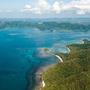 Vista aerea di una costa tropicale con lussureggianti colline verdi che si affacciano su un oceano turchese e isole in lontananza.
