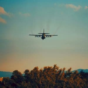 Aereo da trasporto militare visto da dietro in volo a bassa quota sopra le cime degli alberi al tramonto.