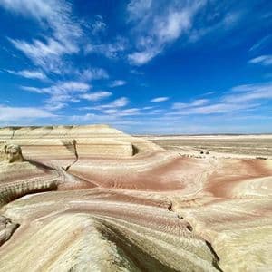 Formazioni rocciose multicolori e stratificate in un vasto paesaggio desertico sotto un cielo azzurro brillante con nuvole, con due veicoli in lontananza.