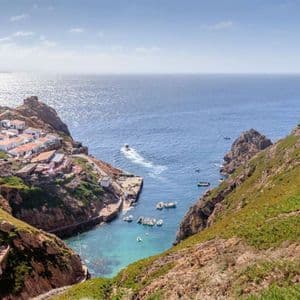 Vista aerea di un borgo costiero con case bianche incastonate in una cala rocciosa e verde. Le barche sono ormeggiate nella baia turchese accanto a un pontile.